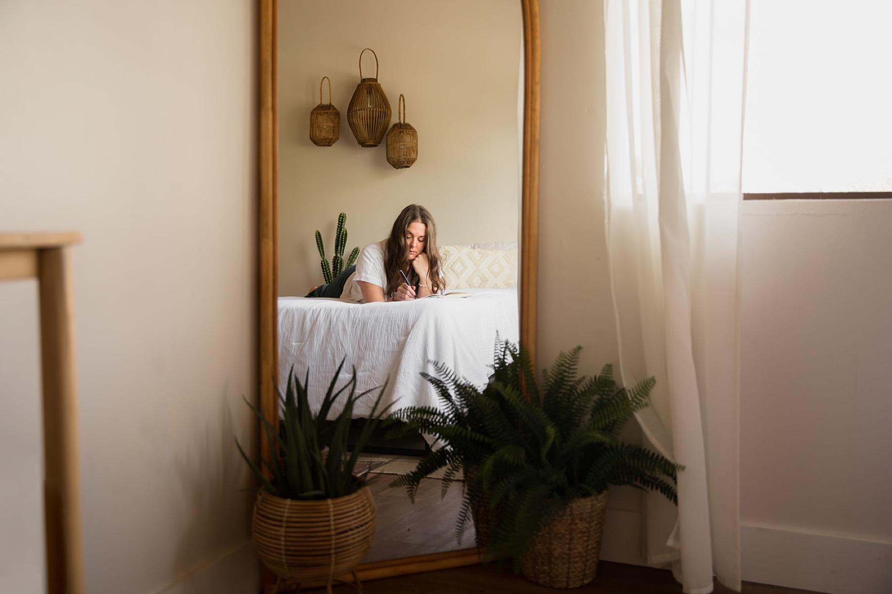 Heather Flynn journaling on a bed, reflected in a tall mirror in a softly lit, plant-filled room